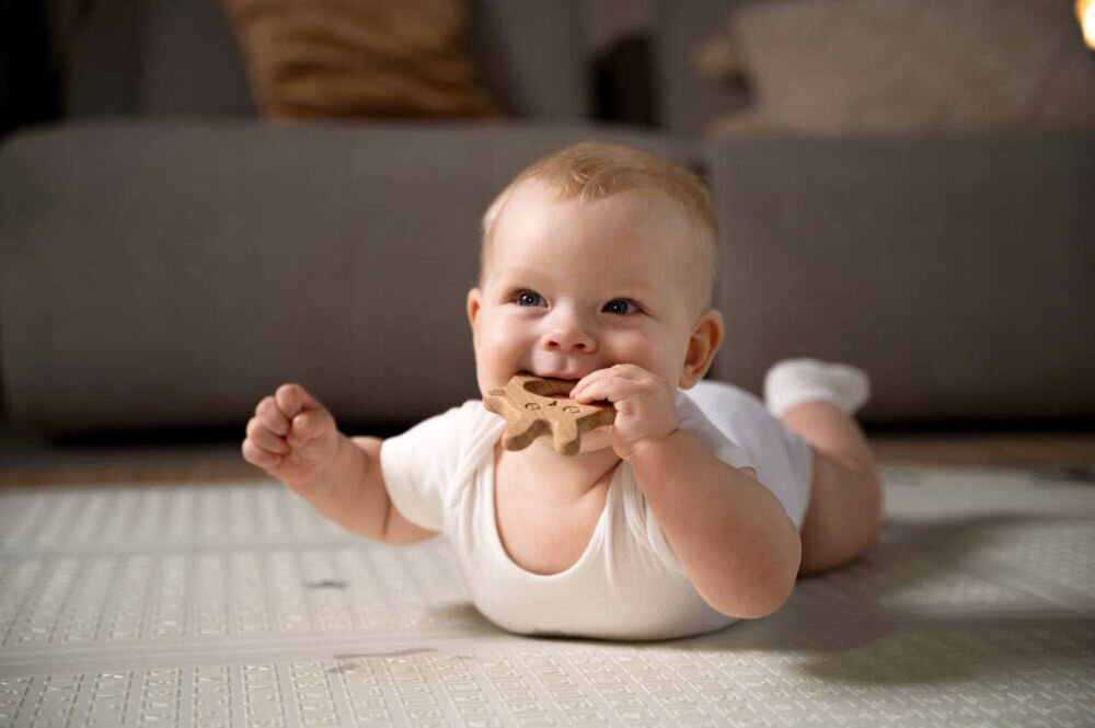 Mata piankowa EXTRALINK 4329 Składana 150 x 200 x 1 cm Niemowlę leżące na brzuszku na miękkiej, szarej macie piankowej Extralink. Dziecko gryzie drewniany gryzak, ćwicząc mięśnie karku w bezpiecznej przestrzeni do zabawy.