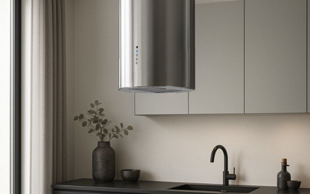 OKAP AKPO WK-12 ISLA BALMERA SLIM Na zdjęciu widać kuchnię z cylindrycznym okapem sufitowym zawieszonym nad blatem. W tle znajdują się jasne szafki, zlew z baterią oraz dekoracje w postaci wazonu z gałązkami i butelek.