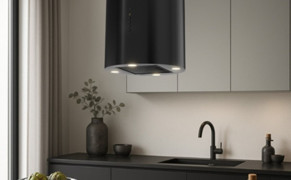 OKAP AKPO WK-12 ISLA BALMERA SLIM Na zdjęciu widać kuchnię z czarnym, cylindrycznym okapem sufitowym zawieszonym nad blatem. W tle znajdują się jasne szafki, zlew z czarną baterią oraz dekoracje w postaci wazonu z gałązkami i butelek.