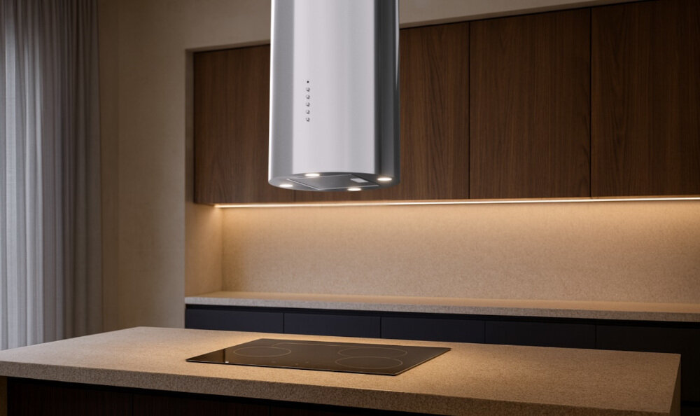 OKAP AKPO WK-4 Isla Balmera Slim Na zdjęciu widać kuchnię z czarnym, cylindrycznym okapem sufitowym zawieszonym nad blatem. W tle znajdują się jasne szafki, zlew z czarną baterią oraz dekoracje w postaci wazonu z gałązkami i butelek.