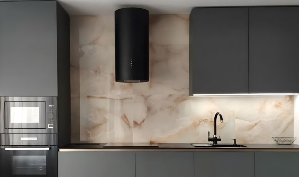 OKAP AKPO WK-12 Balmera WL Slim Na zdjęciu widać kuchnię z czarnym, cylindrycznym okapem sufitowym zawieszonym nad blatem. W tle znajdują się jasne szafki, zlew z czarną baterią oraz dekoracje w postaci wazonu z gałązkami i butelek.