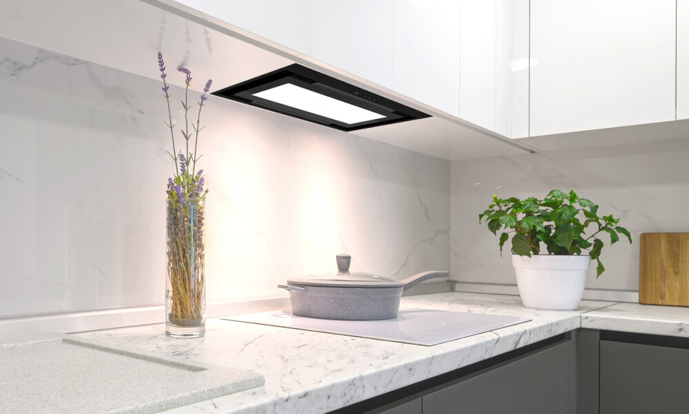 OKAP AKPO WK-12 Evita 60 Widzimy czarny okap sufitowy wbudowany w podwieszany sufit nad jasną strefą kuchenną. Obok znajduje się zielona ściana z rozłożystą rośliną doniczkową, co dodaje wnętrzu świeżości.