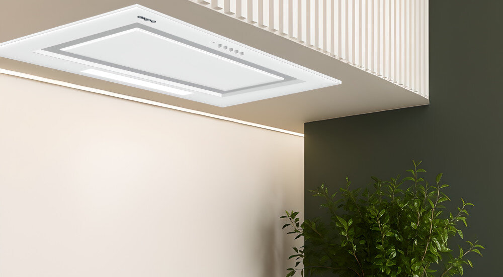 OKAP AKPO WK-7 Fabio 60 Widzimy biały okap sufitowy wbudowany w podwieszany sufit nad jasną strefą kuchenną. Obok znajduje się zielona ściana z rozłożystą rośliną doniczkową, co dodaje wnętrzu świeżości.