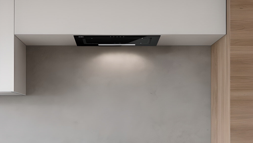 OKAP AKPO WK-7 Fabio 60 Widzimy czarny okap sufitowy wbudowany w podwieszany sufit nad jasną strefą kuchenną. Obok znajduje się zielona ściana z rozłożystą rośliną doniczkową, co dodaje wnętrzu świeżości.