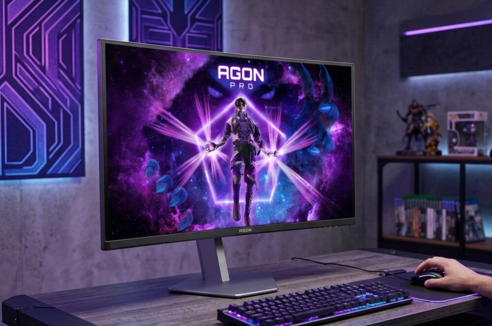 Monitor AOC Agon Pro AG276UZD Monitor stojący na biurku, wyświetlający grafikę z postacią z gry. Obok leży klawiatura oraz ręka na myszce