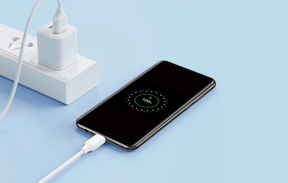 Smartfon podłączony do białego kabla USB, który jest wpięty do listwy zasilającej. Na ekranie telefonu widoczny jest wskaźnik ładowania z napisem \'100%\'.