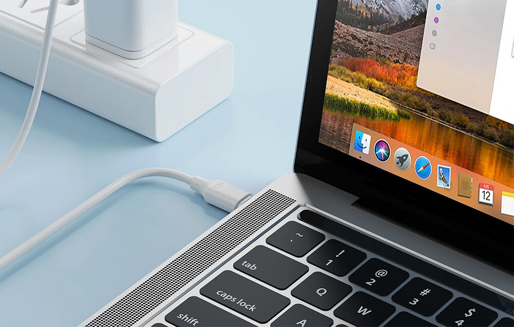 Na obrazie widać laptopa z podłączonym kablem USB typu C, który jest wpięty do białej listwy zasilającej. Na ekranie laptopa widoczne są ikony aplikacji oraz fragment tapety z krajobrazem.