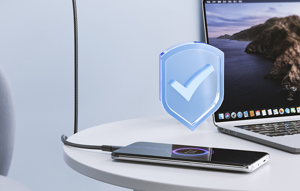 Na białym stole znajduje się smartfon podłączony do laptopa za pomocą czarnego kabla. Obok urządzeń widoczna jest niebieska ikona tarczy z zaznaczeniem.