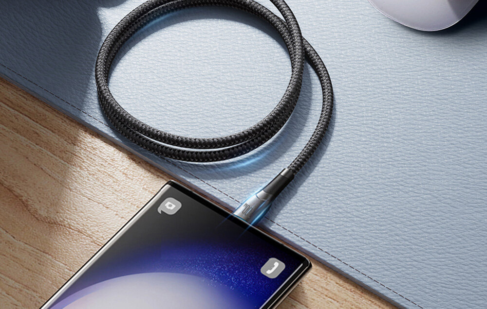 Czarny kabel USB-C podłączony do smartfona leżącego na drewnianej powierzchni obok szarej podkładki. Kabel ma plecioną strukturę i metalowe wykończenie przy złączu.