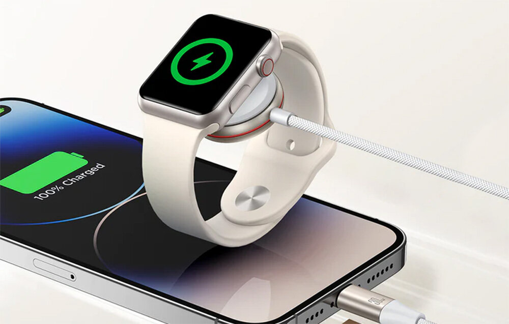 Na obrazie widać zegarek Apple Watch ładowany magnetycznie, umieszczony na białej ładowarce, która jednocześnie ładuje smartfon. Na ekranie zegarka widoczny jest symbol ładowania, a na ekranie telefonu napis \'100% Charged\'.