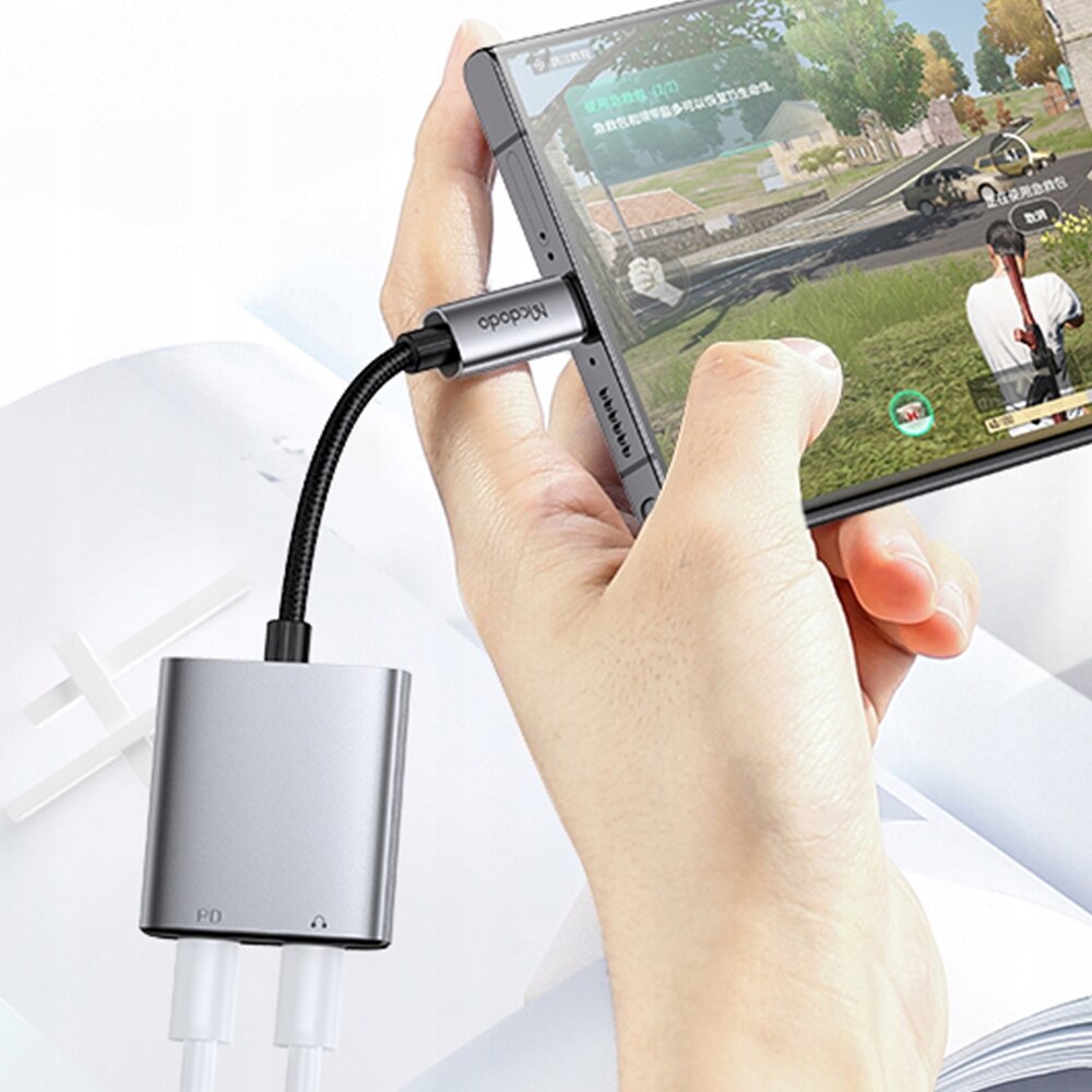 Osoba trzyma smartfon podłączony do adaptera Mcdodo z dwoma portami USB-C. Na ekranie telefonu widoczna jest gra wideo, a adapter jest podłączony do urządzenia za pomocą kabla.