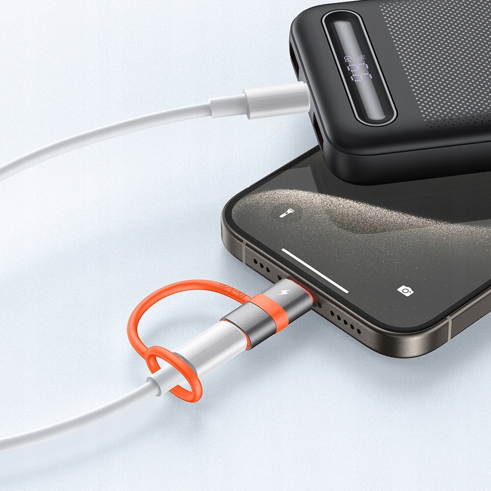 Na obrazie widać smartfon podłączony do powerbanku za pomocą adaptera USB-C do Lightning. Adapter ma srebrno-pomarańczowy design, a kabel jest biały.