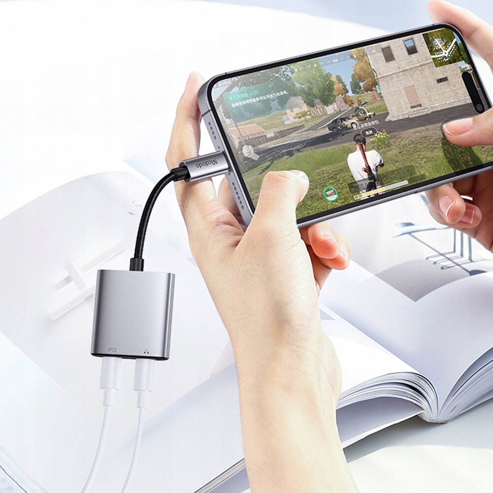 Osoba trzyma smartfon podłączony do adaptera z dwoma kablami Lightning. Na ekranie telefonu widoczna jest gra, a na adapterze znajduje się napis \'Mcdodo\'.