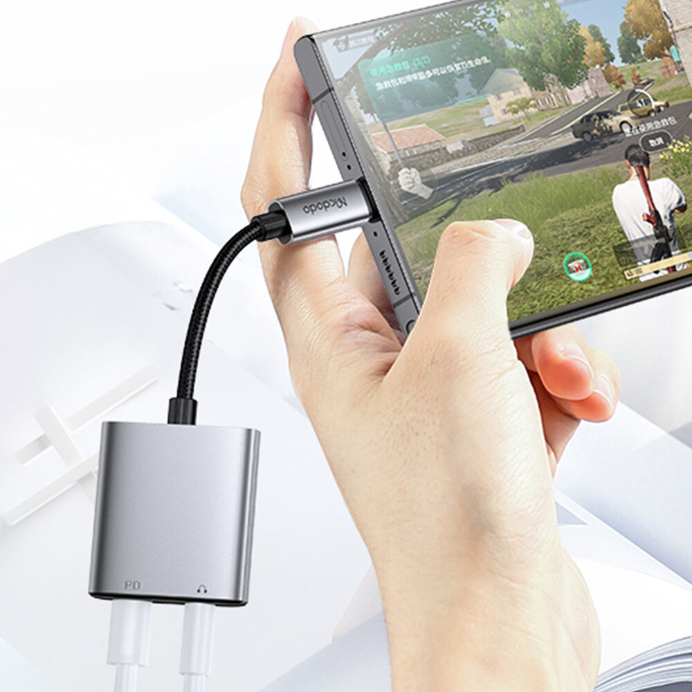 Osoba trzyma smartfon, do którego podłączony jest adapter z logo \'Mcdodo\'. Adapter jest połączony z dwoma białymi kablami, a na ekranie telefonu widoczna jest gra.