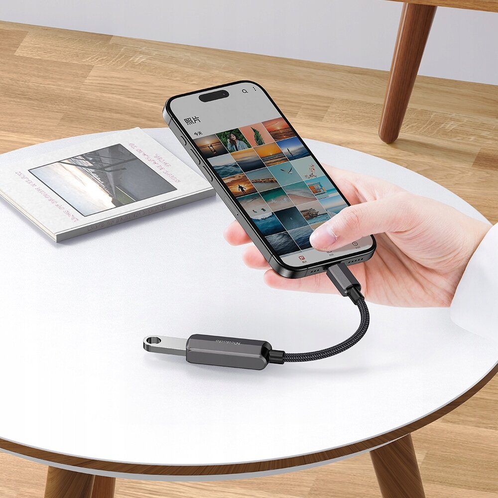 Na okrągłym stole leży telefon podłączony do adaptera USB-C na USB-A marki Mcdodo, obok znajduje się otwarta książka z widocznym zdjęciem na okładce.