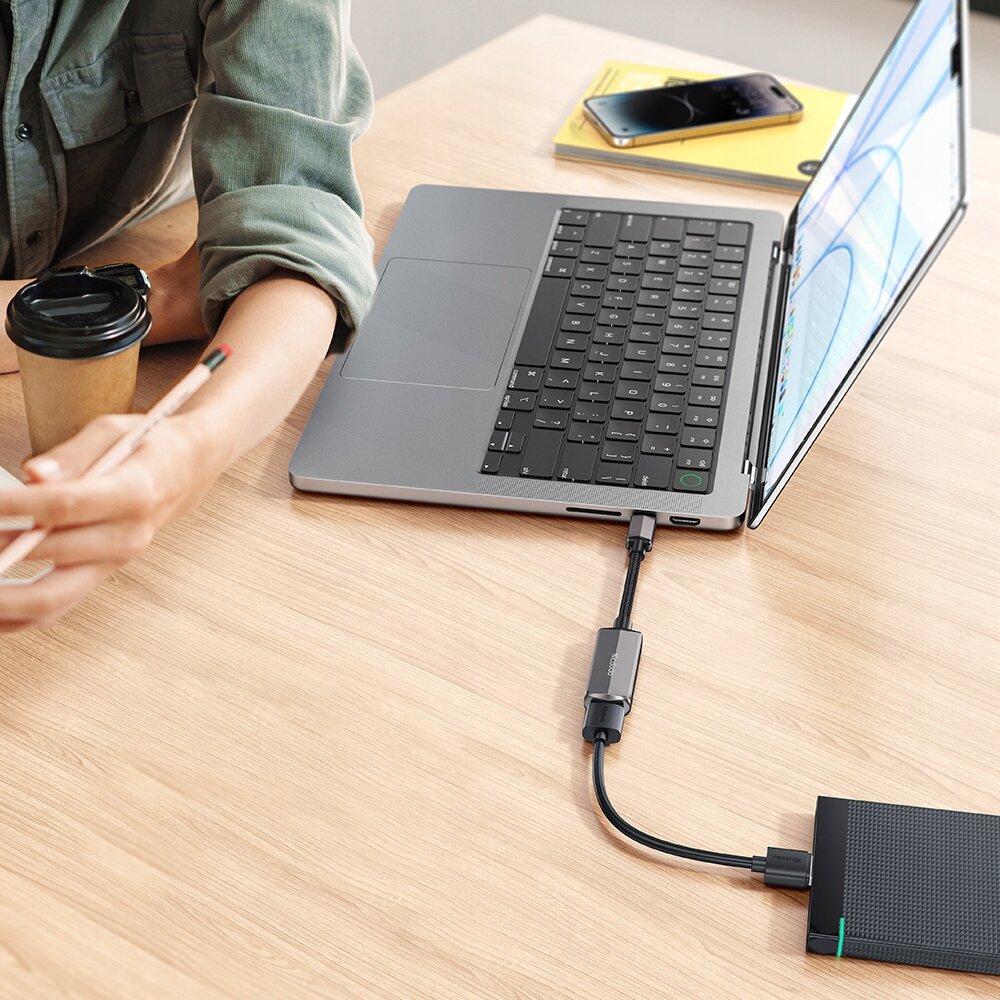 Osoba siedzi przy biurku z laptopem, do którego podłączony jest adapter USB-C na USB-A. Obok znajduje się kubek z napojem i smartfon leżący na notatniku.