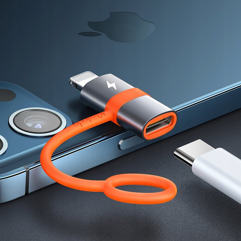 Na zdjęciu znajduje się adapter USB-C do Lightning z pomarańczowym kablem, obok smartfona z widocznym logo w kształcie jabłka. Na adapterze widoczny jest napis \'Mcdodo\'.