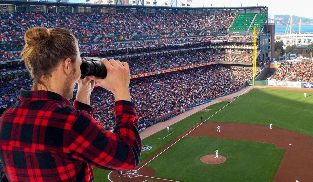 Osoba w czerwono-czarnej koszuli patrzy przez lornetkę na stadionie podczas meczu baseballowego. W tle widać pełne trybuny i boisko z zawodnikami.