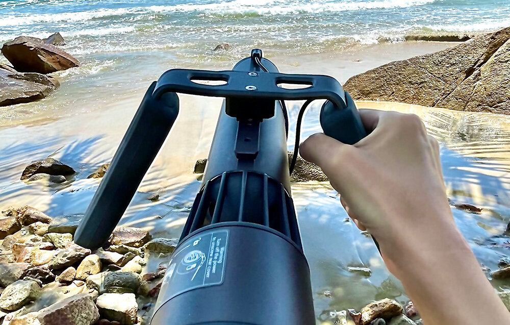 Uchwyt do skutera WAYDOO 4010000050 plaża osoba trzyma skuter podwodny za pomocą uchwytu w tle woda podnosi komfort wypraw stabilne i pewne trzymanie urządzenia pasuje do naturalnego ułożenia dłoni swobodne manewrowanie