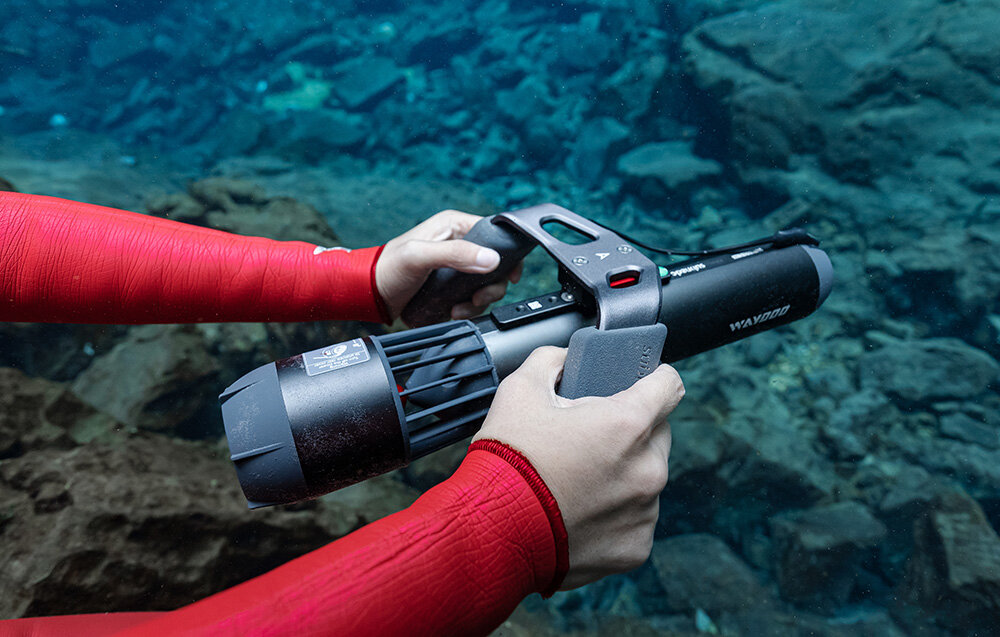 Uchwyt do skutera WAYDOO 4010000050 akwen wodny osoba trzyma skuter podwodny za pomocą uchwytu w tle kamienie ergonomiczny kształt uchwytu pewne prowadzenie skutera stabilność ułatwia precyzyjne kontrolowanie kierunku wykonywanie płynnych manewrów