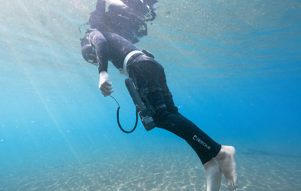Kontroler na palec do skutera podwodnego WAYDOO 4010000053 akwen wodny nurek zbliżenie na nogi nurka nurek w dłoni trzyma kontroler na nogach ma przymocowane napędy skutera wodnego do skutera Waydoo Subnado pewne trzymanie urządzenia naturalne ułożenie ręki swobodne manewrowanie za pomocą kciuka kierowanie napędem staje się proste i bardzo naturalne
