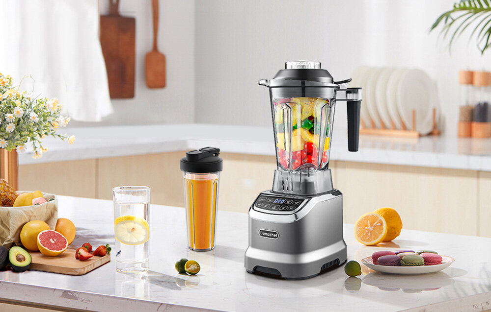 Na blacie kuchennym znajduje się blender kielichowy z kolorowymi owocami w środku oraz szklanka z napojem i butelka z sokiem. Obok leżą owoce, kwiaty i makaroniki, a na blenderze widoczne jest logo \'amzchef\'.
