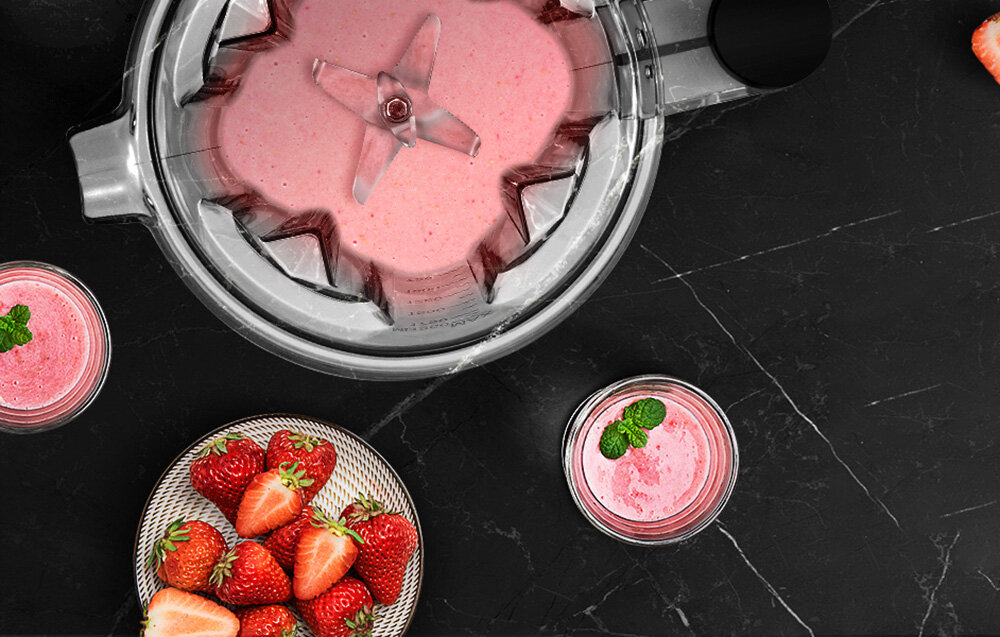 Na czarnym blacie znajduje się blender kielichowy z różowym koktajlem w środku, obok stoją dwa szklanki z podobnym napojem. Na talerzu leżą świeże truskawki.
