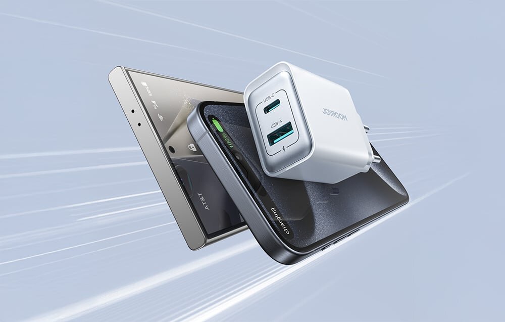 Na obrazie widoczna jest biała ładowarka sieciowa z dwoma portami USB oznaczonymi jako USB-A i USB-C oraz napisem \'JOYROOM\'. W tle znajdują się trzy smartfony ułożone jeden na drugim.