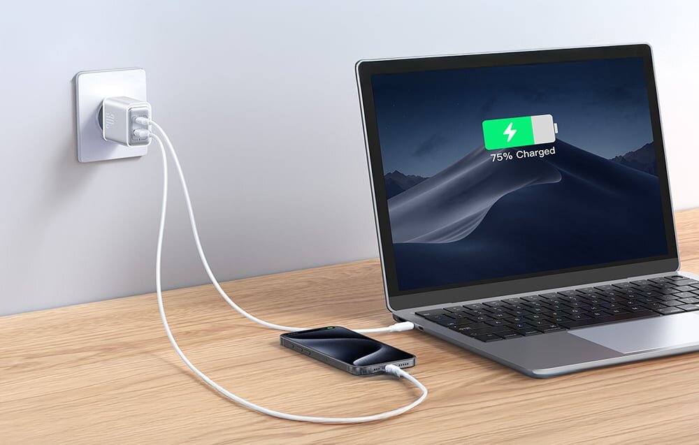 Na drewnianym blacie stoi laptop z ekranem pokazującym ikonę baterii z napisem \'75% Charged\'. Obok leży smartfon podłączony do ładowarki sieciowej w gniazdku, z dwoma kablami USB.