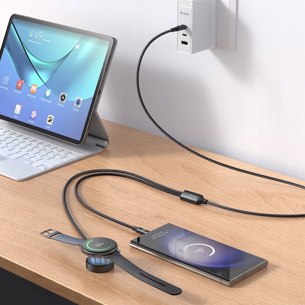 Na drewnianym blacie znajduje się tablet z klawiaturą, telefon oraz zegarek ładowane za pomocą kabla USB-C podłączonego do ładowarki w gniazdku. Na ładowarce widoczne jest logo \'Mcdodo\'.