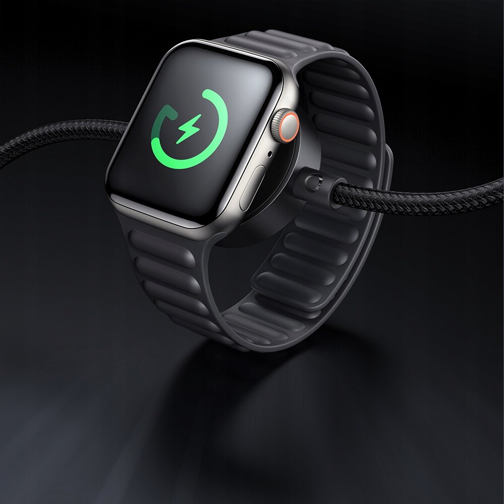 Na obrazie widoczny jest smartwatch z czarnym paskiem, na ekranie którego wyświetla się zielona ikona ładowania. Do zegarka podłączony jest czarny kabel z plecionym wzorem.