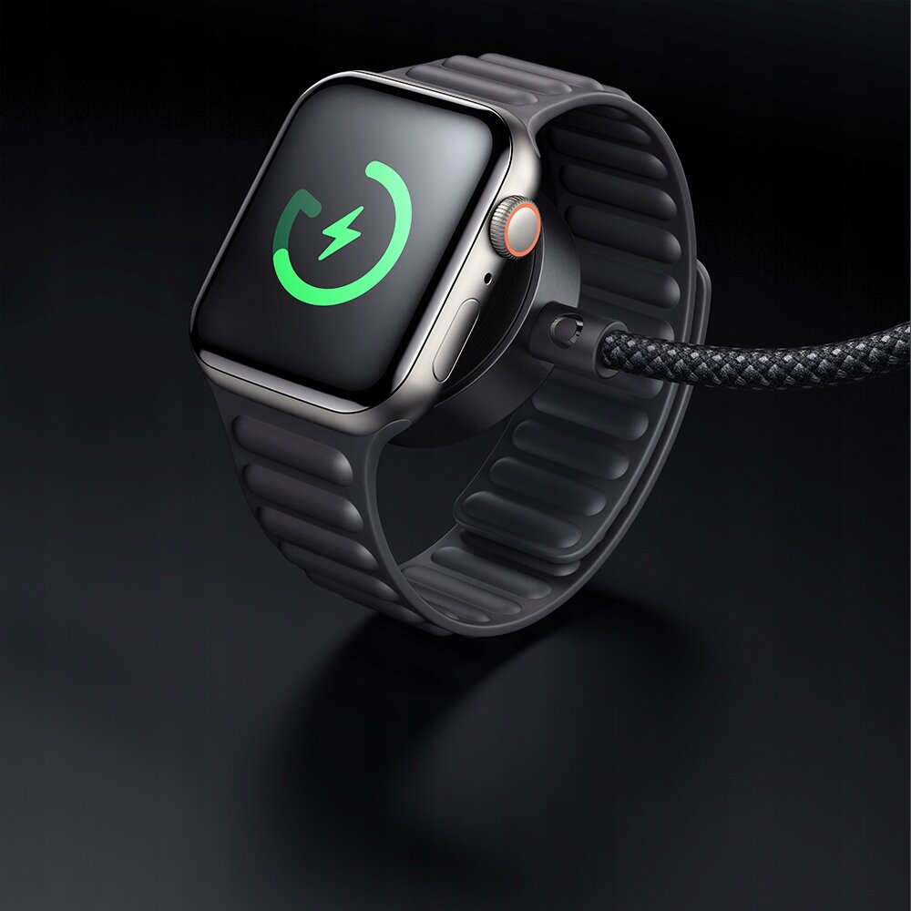 Na obrazie widoczny jest smartwatch z czarnym paskiem, na którego ekranie wyświetla się zielona ikona ładowania. Do zegarka podłączony jest pleciony kabel.