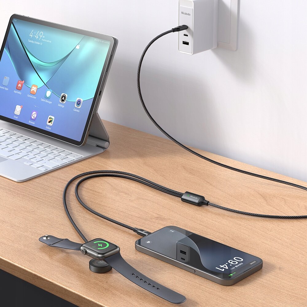 Na drewnianym blacie znajduje się tablet z klawiaturą, smartfon oraz zegarek na ładowarce indukcyjnej. Kabel z napisem \'Mcdodo\' podłączony jest do gniazdka ściennego.