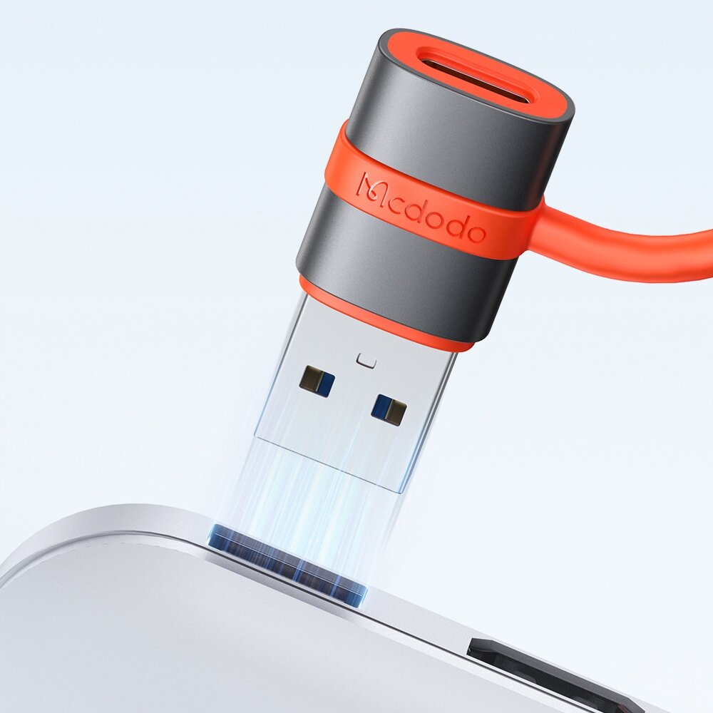 Na obrazie widoczny jest adapter USB z logo \'Mcdodo\', w kolorze szaro-pomarańczowym, podłączany do portu USB w urządzeniu. Adapter ma kształt prostokątny z okrągłym zakończeniem i pomarańczowym kablem.