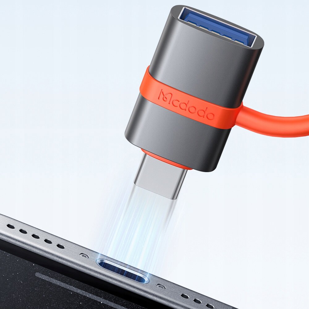 Na obrazie widoczny jest adapter USB z logo \'Mcdodo\' na obudowie, podłączany do portu urządzenia. Adapter ma metaliczną obudowę z pomarańczowym elementem i niebieskim wnętrzem złącza USB.