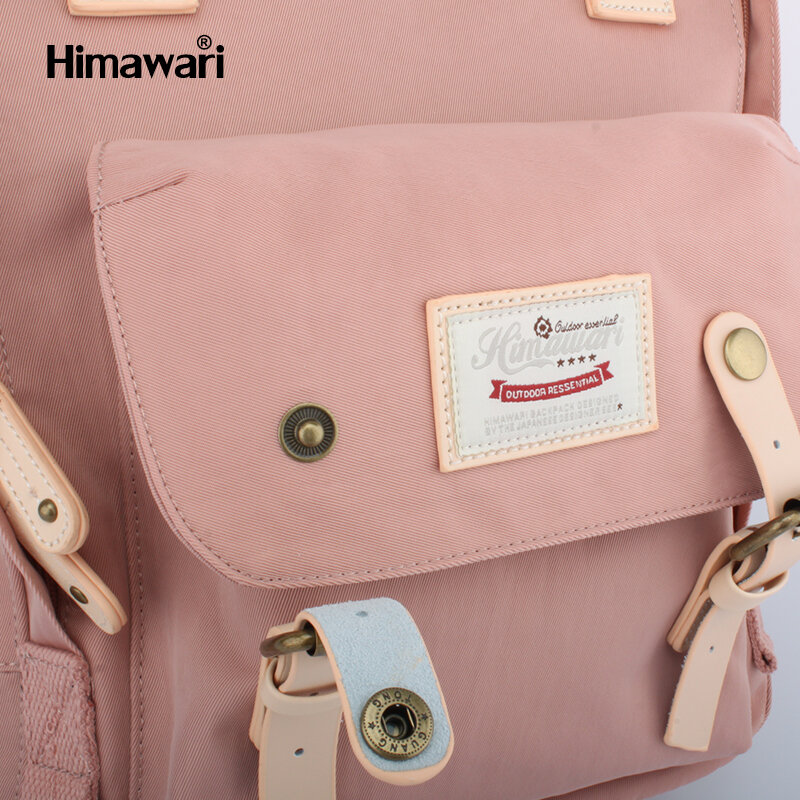 Różowy plecak z klapą i kieszenią z przodu, ozdobiony metalowymi zatrzaskami i skórzanymi paskami. Na kieszeni znajduje się naszywka z napisem \'Himawari Outdoor Essential\'.