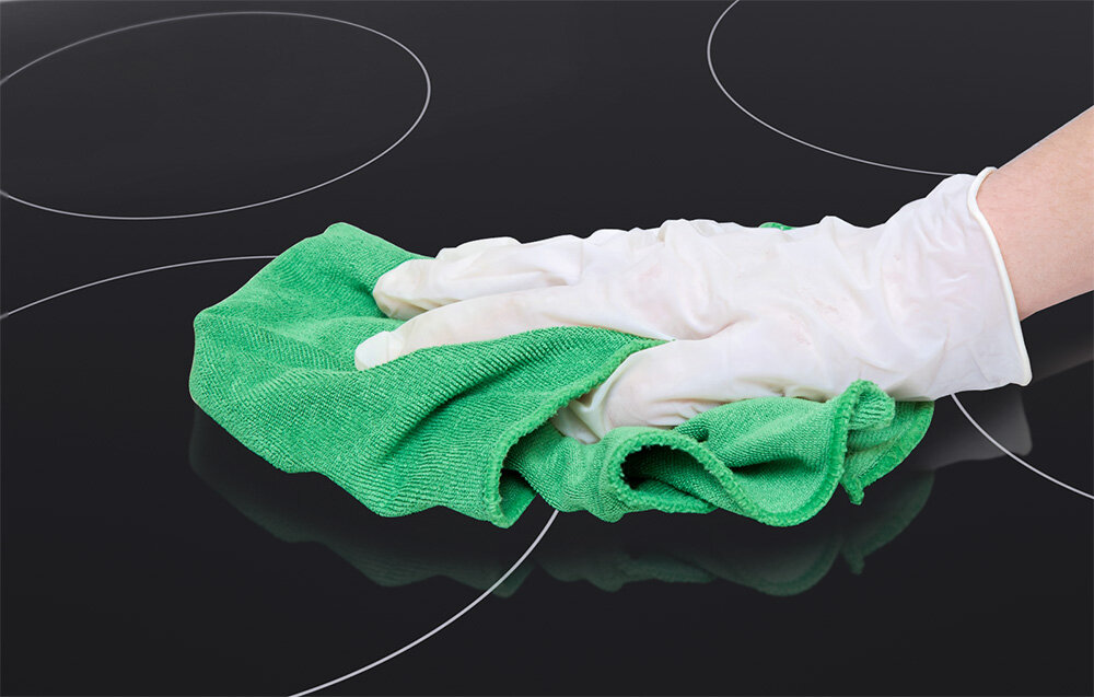 PŁYTA CERAMICZNA ISEASY T4-04 Powierzchnia płyty jest przecierana zieloną ściereczką przez osobę w białej rękawiczce. Zdjęcie pokazuje prostotę czyszczenia gładkiej tafli szkło hartowane łatwe czyszczenie powierzchnia