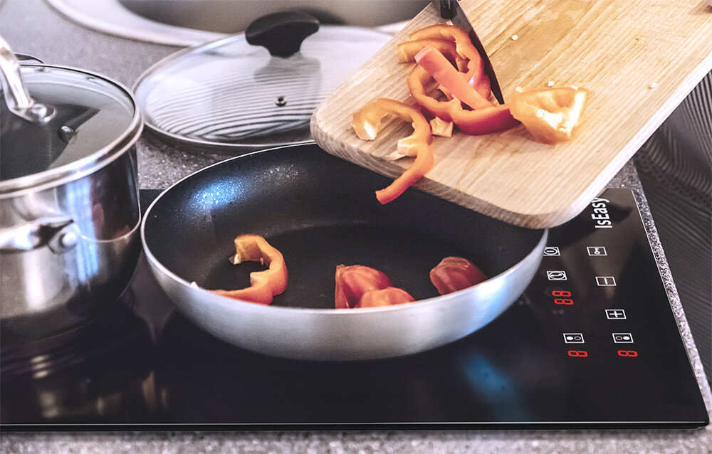 Płyta ceramiczna ISEASY T2V-01M Pokrojone warzywa są zsuwane z deski na patelnię stojącą na płycie grzewczej. Obok widać garnek z pokrywką, tworzący kuchenną scenę przygotowywania posiłku płyta ceramiczna 2 pola 1800W 1200W
