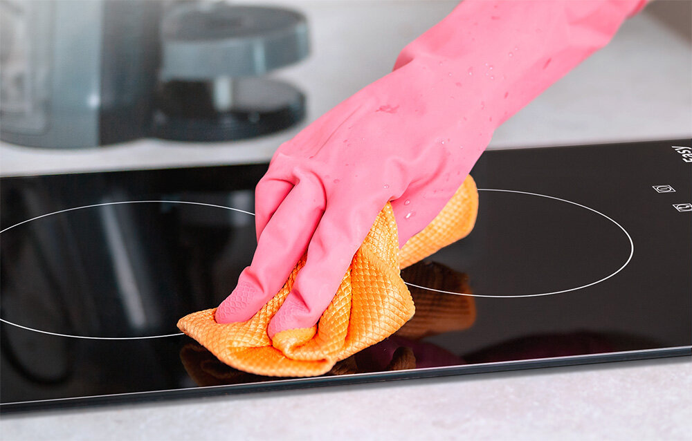 Płyta ceramiczna ISEASY T2V-01M Dłoń w różowej rękawiczce przeciera płytę grzewczą pomarańczową ściereczką. Zdjęcie podkreśla prostotę utrzymania powierzchni w czystości szklana powierzchnia łatwe czyszczenie płyta bez szorowania
