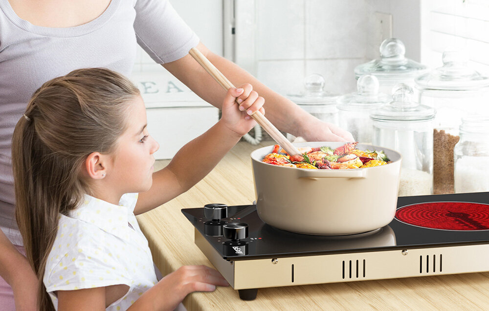 Płyta ceramiczna ISEASY LT2V-30 Osoby gotują posiłek w dużym, jasnym garnku ustawionym na płycie, mieszając zawartość drewnianą łopatką. Blat otaczają słoiki i kuchenne dodatki, tworząc ciepłą atmosferę gotowania płyta ceramiczna 2 pola pokrętła