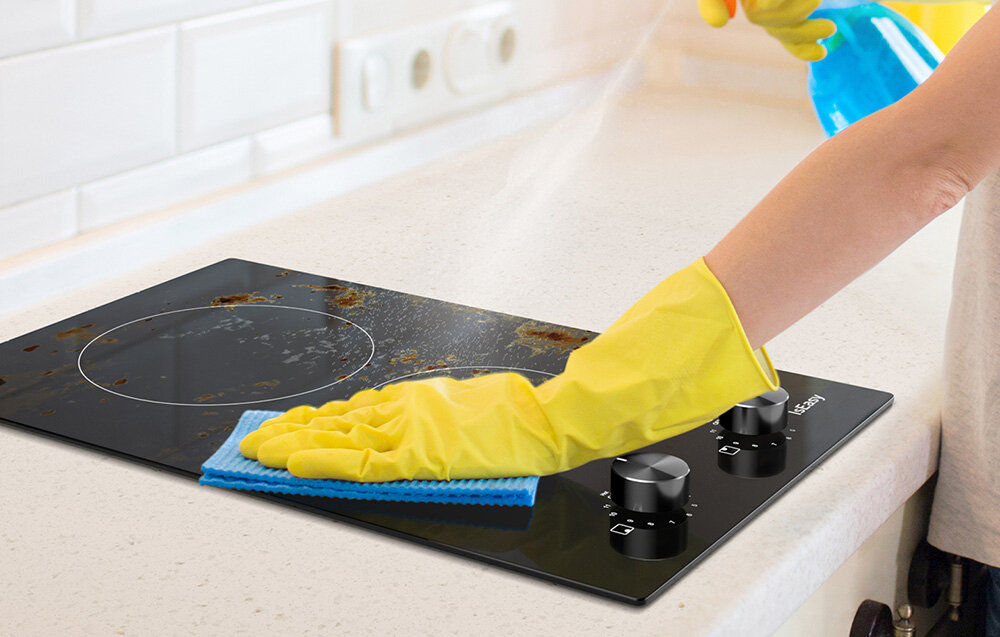 Płyta ceramiczna ISEASY LT2V-30 Ręka w żółtej rękawicy czyści płytę grzewczą z zabrudzeń, używając niebieskiej ściereczki. W tle widać spryskiwacz z detergentem, a płyta jest częściowo pokryta zaschniętymi plamami wskaźnik ciepła automatyczne wyłączanie bezpieczeństwo