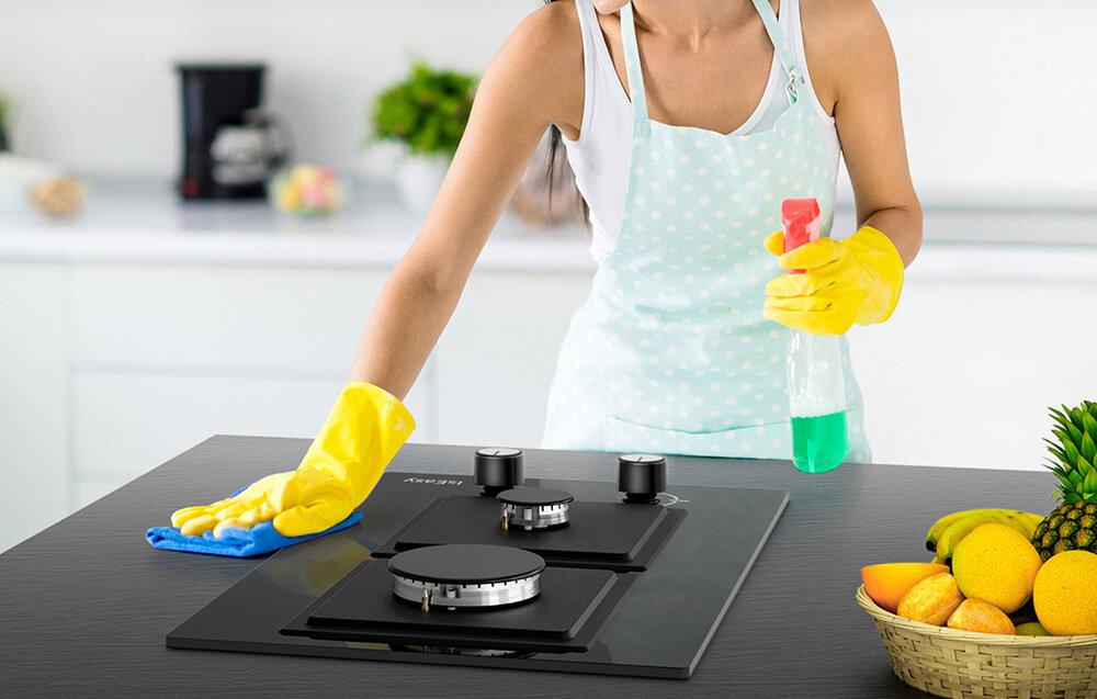 PŁYTA GAZOWA ISEASY MGBG-312S2C Osoba czyści powierzchnię płyty gazowej miękką ściereczką, a obok stoją świeże owoce. Detale palników i pokręteł są dobrze widoczne na czarnym panelu żeliwne ruszty szklana płyta łatwe czyszczenie odporność na temperaturę