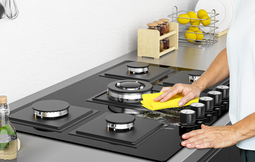 PŁYTA GAZOWA ISEASY MGBG-775S2 Osoba czyści powierzchnię płyty gazowej miękką ściereczką, przesuwając ją wzdłuż panelu. Widoczne są także elementy palników w pełnym układzie pięciu miejsc szklana powierzchnia łatwe czyszczenie trwałe pokrętła solidne palniki