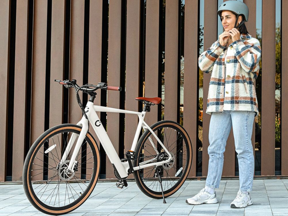 Rower elektryczny CECOTEC Sprint M20 28 cali Biały chodnik ściana budynku rower oparty na stopce obok stoi osoba zakłada kask składana stopka postój bezpieczny i bezproblemowy wytrzymała nóżka w centralnym punkcie ciężkości