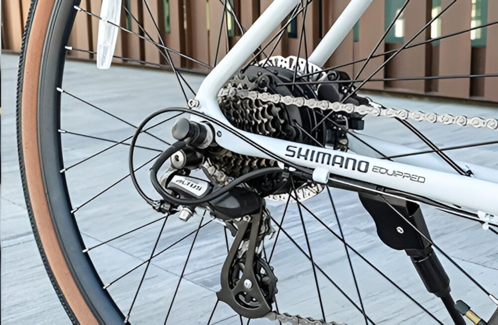 Rower elektryczny CECOTEC Sprint M20 28 cali Biały chodnik ściana budynku zbliżenie na przerzutkę manetki SHIMANO typu SL-M315 8 biegów tylna przerzutka SHIMANO Altus 8S technologię SHADOW przerzutka jest schowana i mniej narażona na uszkodzenia duża wytrzymałość precyzyjne działanie zmiana biegów jest płynna i bezproblemowa