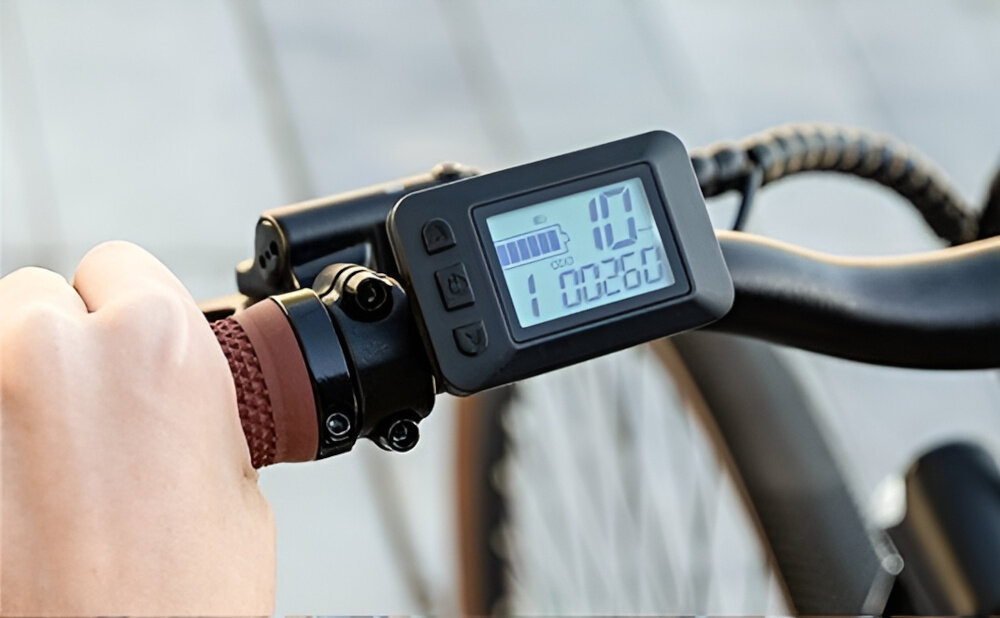 Rower elektryczny CECOTEC Sprint M20 28 cali Szary chodnik zbliżenie na wyświetlacz osoba trzyma dłoń na kierownicy na wyświetlaczu różne liczby aluminiowa kierownica antypoślizgowy materiał pewny chwyt komputer pokładowy LCD