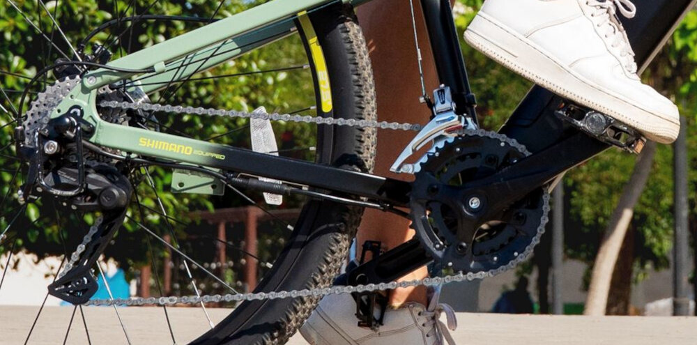 Rower elektryczny CECOTEC Millor U18 27.5 cala Zielony miasto drzewa zbliżenie na korbę i wolnobieg osoba opiera stopę na pedale lekka i aluminiowa konstrukcja korby marki SHIMANO wolnobieg SHIMANO typu AMFTZ5007428 solidny wytrzyma obciążenie nawet w trudnych warunkach wytrzyma bardzo duży nacisk na pedały
