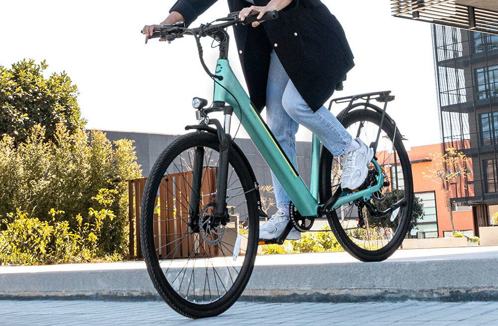 Rower elektryczny CECOTEC Urban D18 28 cali Niebieski chodnik budynki drzewa niebo osoba jedzie na rowerze zjeżdża z krawężnika rower typu hardtail przedni amortyzator marki Orbi Shocks typu XCE28 sprężyna stalowa skutecznie tłumi wstrząsy regulacja wstępnego napięcia