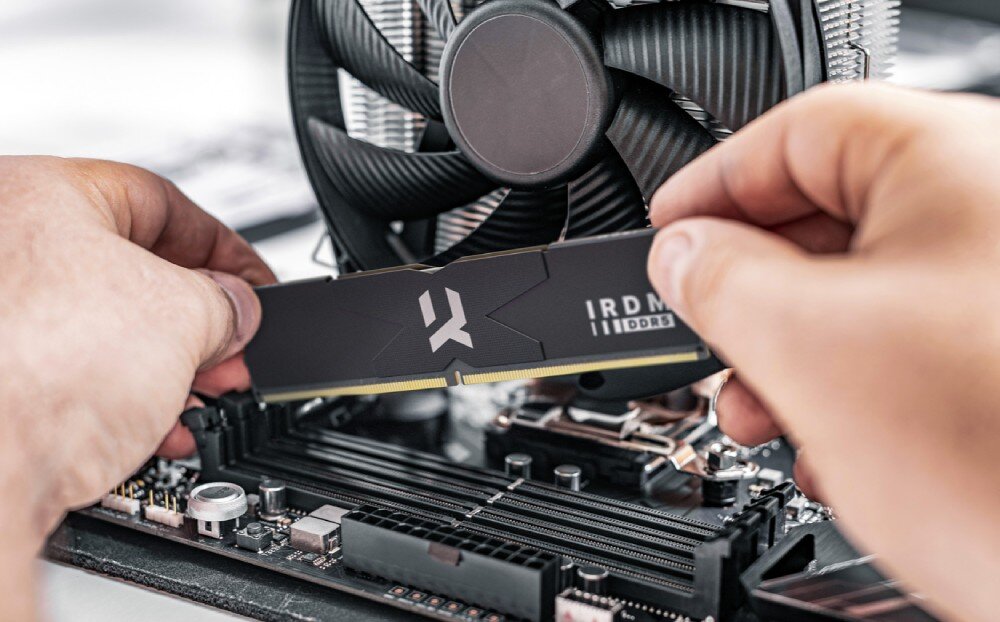 Pamięć RAM GOODRAM IRDM DDR5 Dłonie instalujące czarną kość pamięci RAM w gnieździe płyty głównej komputera obok dużego wentylatora procesora, pojemność, taktowanie, opóźnienia, konstrukcja