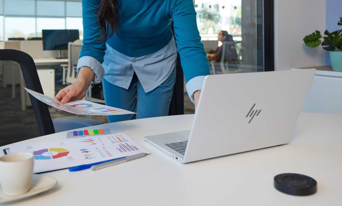 Osoba w niebieskim stroju pochyla się nad białym biurkiem, na którym leży otwarty srebrny laptop HP z widocznym logo, wokół rozłożone są kolorowe wydruki z wykresami, filiżanka kawy i długopis; w tle biuro z rośliną doniczkową. - Opis serii HP EliteBook X G1a